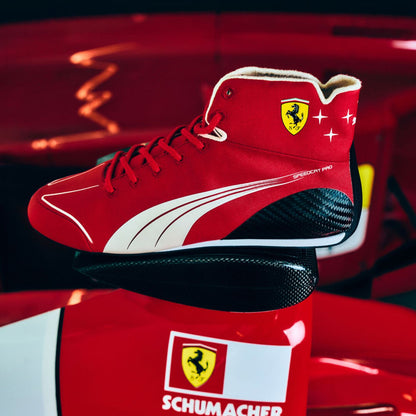 A red Puma Scuderia Ferrari high-top sneaker with white Puma stripes and a yellow Ferrari shield logo, displayed on top of a red and white Ferrari box in a dark red studio setting.