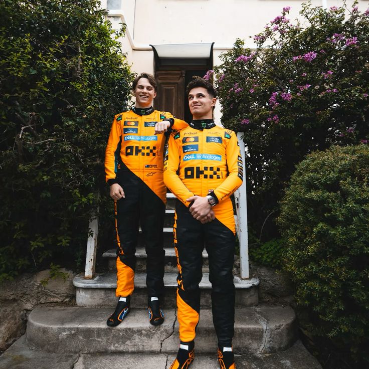 Lando Norris and Oscar Piastri in a 2025 McLaren orange F1 racing suit standing on steps outdoors.