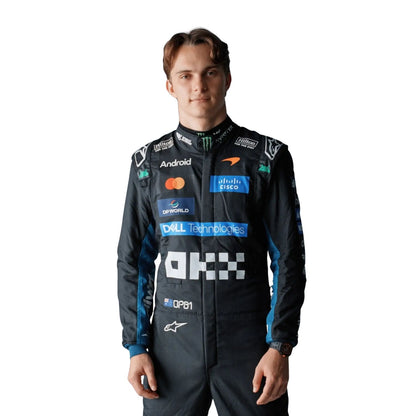 A Oscar Piastri wearing a dark blue and black Alpinestars racing suit with various sponsor logos, including Mastercard, Android, and Dell Technologies, standing against a solid white background.