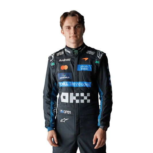 A Oscar Piastri wearing a dark blue and black Alpinestars racing suit with various sponsor logos, including Mastercard, Android, and Dell Technologies, standing against a solid white background.
