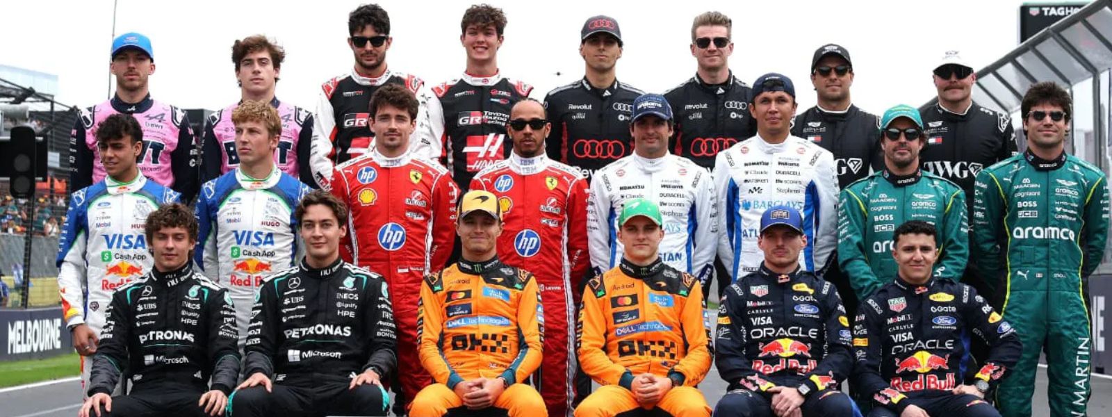 A group photo of Formula 1 racing drivers in their team uniforms, with sponsor logos visible on their suits, set against a racetrack background.